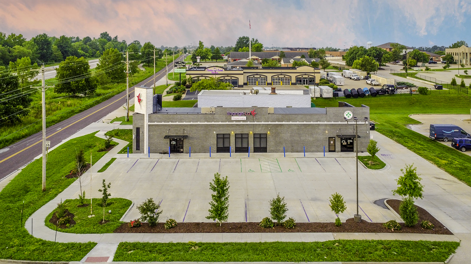 A modern commercial building with a large empty parking lot surrounded by greenery and nearby roads